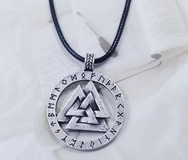 A white background featuring a pad of paper and a pen hosts a viking style necklace. A black cord is just seen as the chain and the main design is a valknut trianglular shape in the middle of a circle of runes which are carved in black.