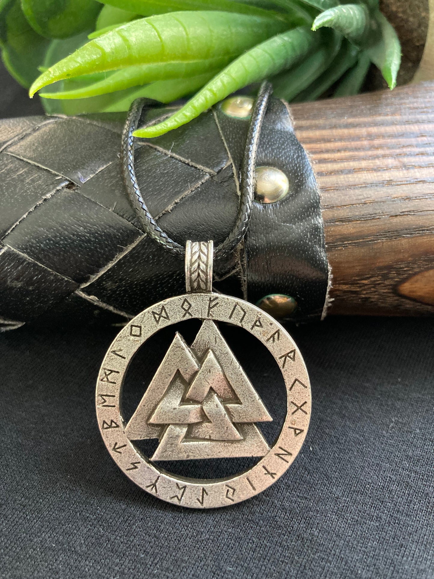 A detailed view of a stainless steel round pendant is on show leaning against an axe handle. The Main design is 3 interlocking triangles as a cut out piece inside a circle of runic letters which are engraved in black around the outside. 