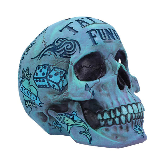 A large blue skull is on a plain background. There are various pictures etched around it as if it has been tattooed. You can see a pair of dice, a shark, a heart and tribal type patterns. The jaw slightly sits forward and the eyes and nose are hollowed out.