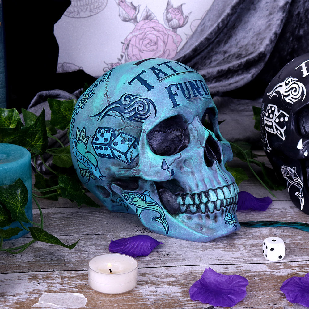 A large skull sits on a table surrounded by petals, a candle and a plant. You can see the stunning detailing of the facial features as well as some of the images made to look like tattoos. 