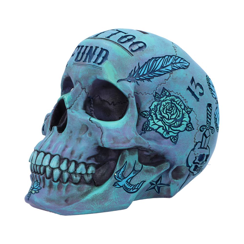 A side view of a blue skull figurine. The cheekbones are sunken in and the eye sockets hollowed out. A wide grimace is on display and there are images etched onto the cranium made to look like tattoos. These include a feather, a rose and a bird.