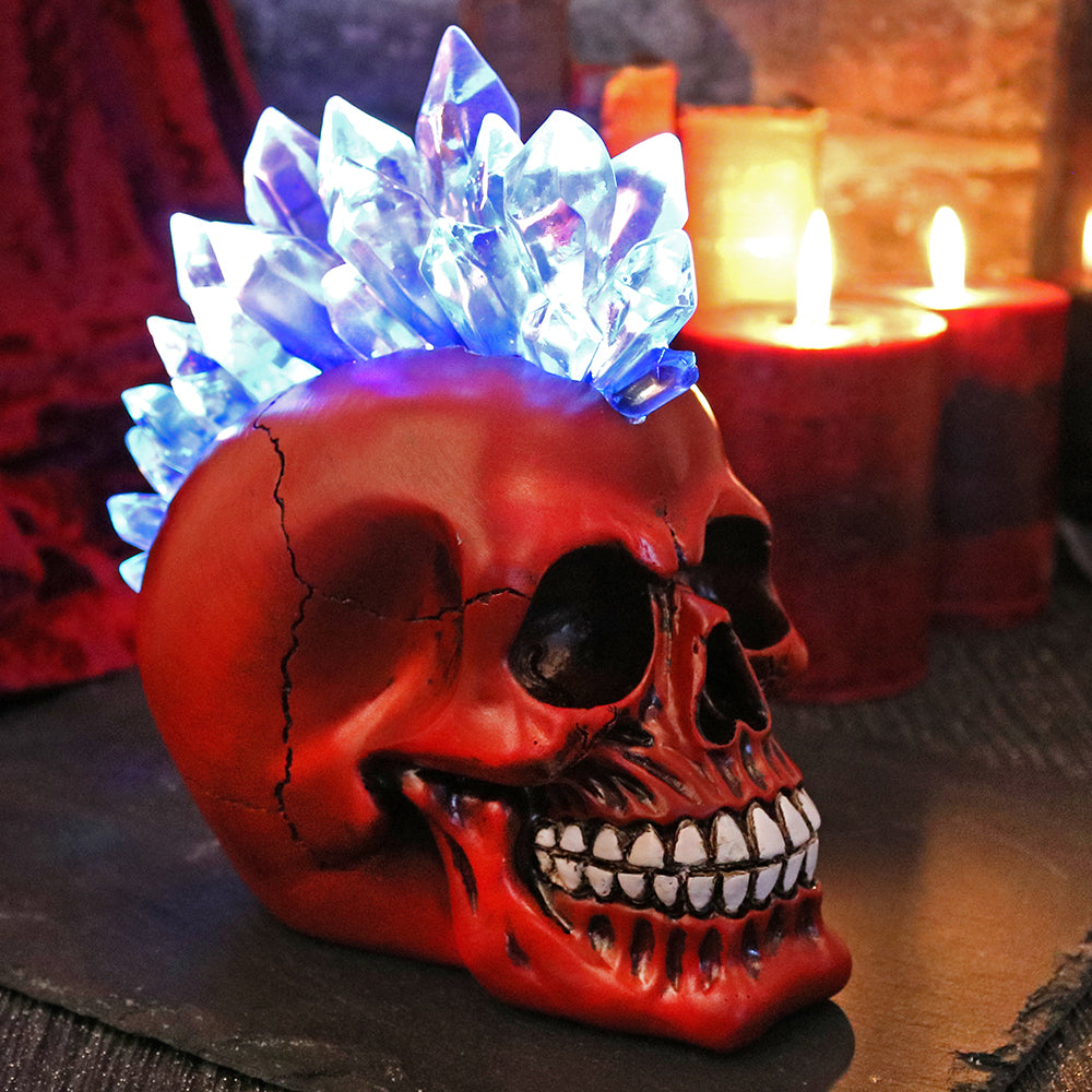 On a surface with candles in the background sits a red skull figure. You can see the light blue crystal looking mohawk is lit up and is shining. The jaw is detailed with sunken in bits around the mouth and teeth. 