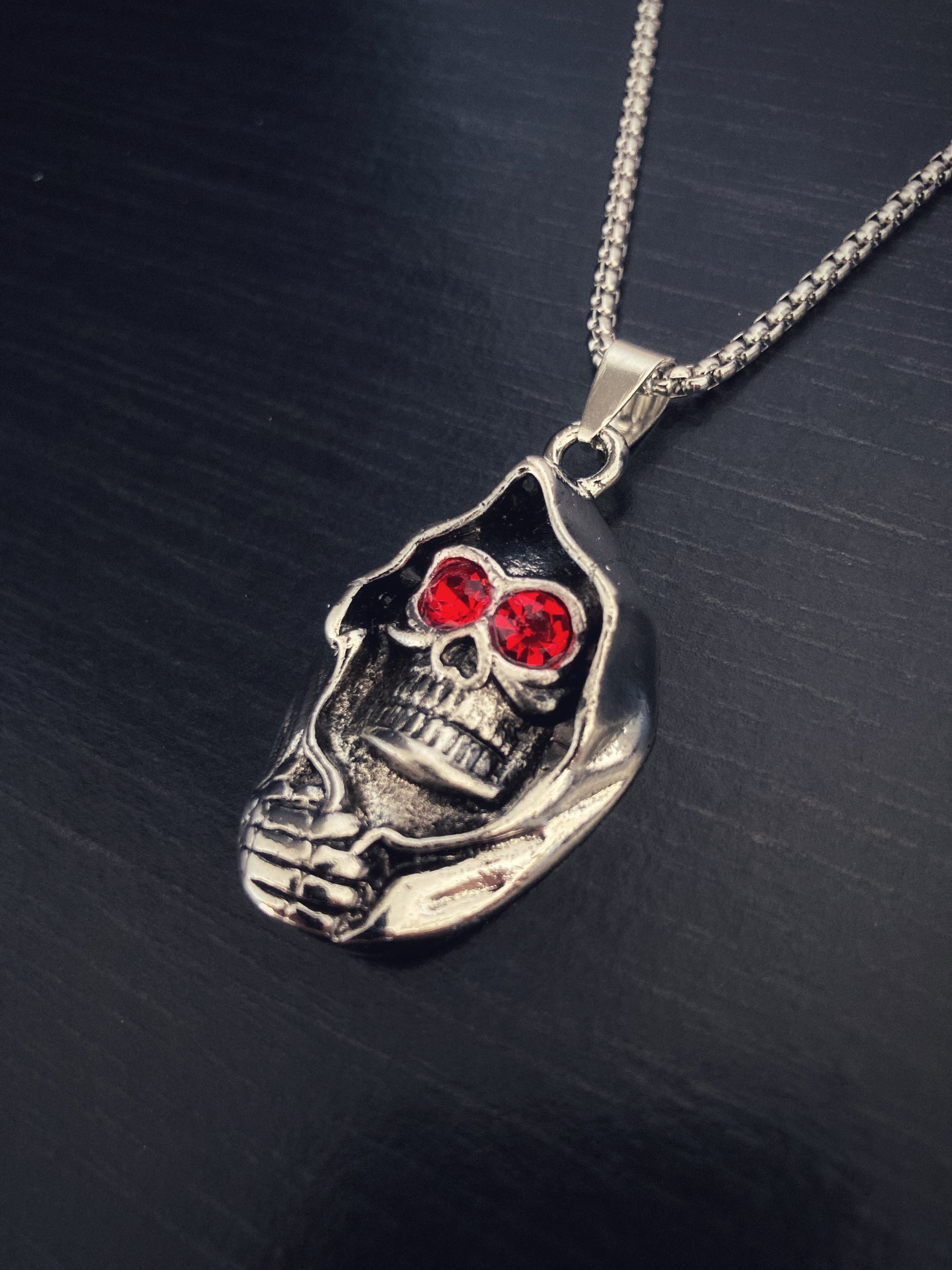 A skull pendant sits on a black surface, it is attached to a sturdy chain necklace sturdy. The item is silver coloured and the skull is cloaked with a creepy hand holding it in place. The eyse have been hollowed out and replaced with red stones.