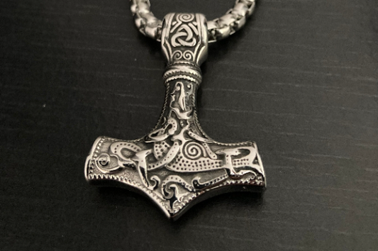 A solid looking pendant in the shape of Thor's hammer sits on a black wooden background. This is a close up of the detailing of the main body of the item which features an intricate pattern which you can also feel when you touch it. 
