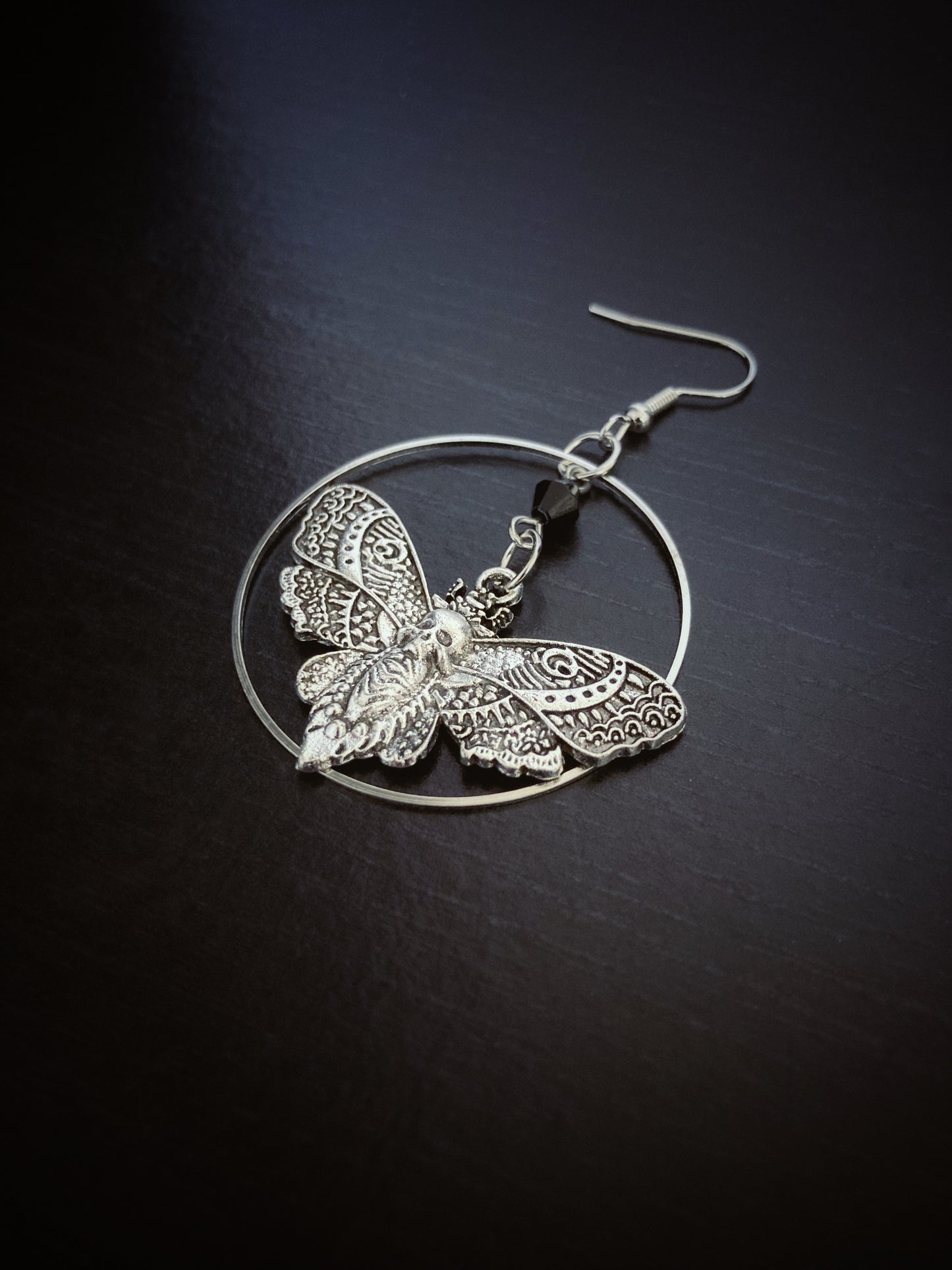 On a black surface sits a singular earring which has a large thin hoop and a death moth nestled inside this. It is silver in colour and incredibly detailed with patterns all over the moth itself.