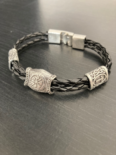 A black leather knotted style band sits on a dark surface. There are 3 silver coloured runes in different styles at intervals on the band. 