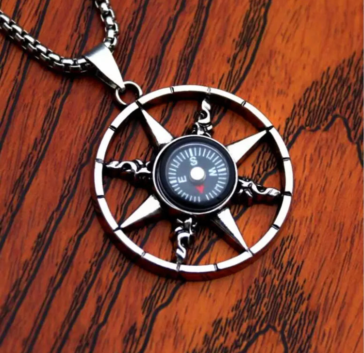 On a brown wooden surface sits a circular compass pendant. There is an outer ring which is narrow and then 8 points touching this with a full working round compass in the middle. It is attached to a sturdy chain for wearing and its silver in colour.