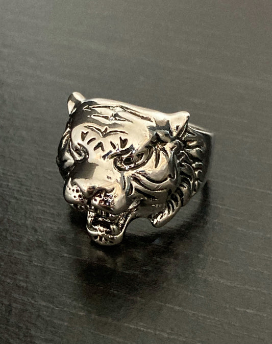 A front view of a detailed tiger head ring that sits upon a black grainy surface. It is ferocious looking with its mouth gaping open and the whole item is solid and weighty.