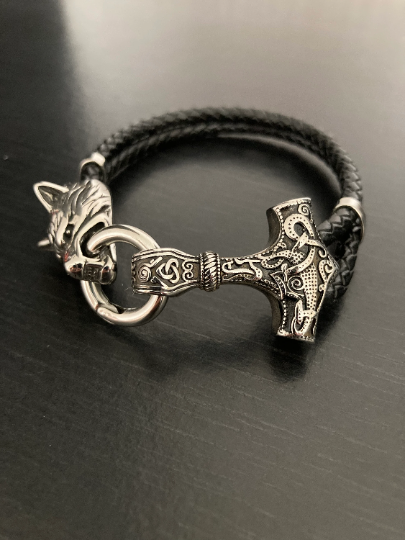 Upon a black background sits a bracelet that has two black leather bands attached to a Thor style hammer one end and a fenrir wolf the other. There is a ring in the wolfs mouth and the other side of the ring is attached to the hammer. 