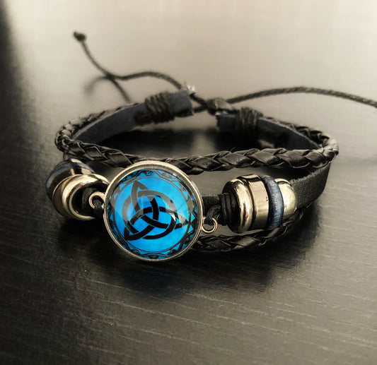 A bracelet that is made up of multi layered bands sits on a black surface. There is a round main feature that has three interlocked triangles in black on a turquiose background. Coming off of that are metal beads and then a plain black leather band with knotted black bands either side of it.