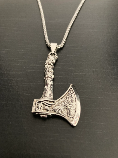 A Silver coloured viking style axe necklace is on a black background. The pendant has an intricate design egfraved on the axe head which is a wolf. The item is attached to a chain which is also silver in colour and is stainless steel