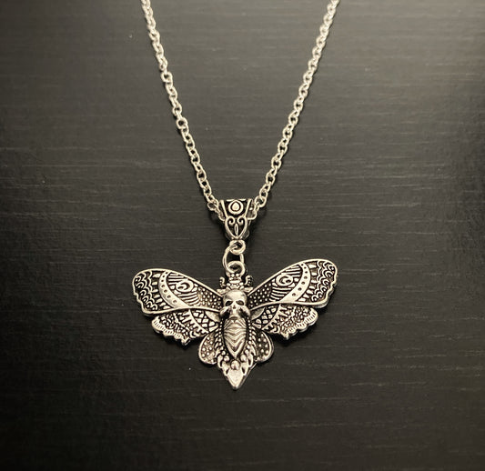 On a black grainy background is a pendant in the shape of a death moth which is attached to a sturdy looking chain. Both are silver in colour and made of stainless steel and the wings of the moth feature an intricate design.