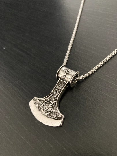 A silver coloured necklace chain sits on a grainy black surface with a pendant in the shape of an axes head attached to it. This has an engraving of a black sun symbol om it as well as a very detailed pattern surrounding that