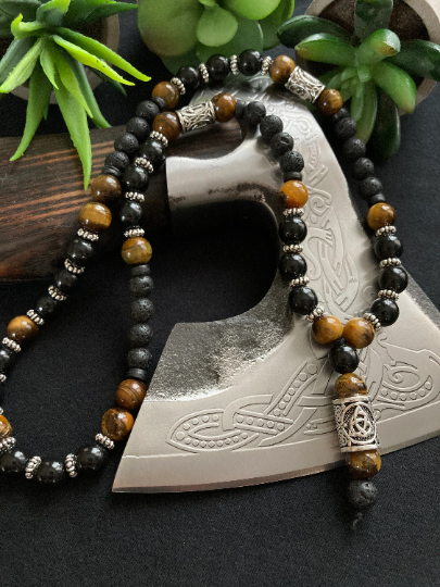 A stunning beaded necklace is draped over an axe. With a section that hangs down at the front and the rest of the item featuring metal cylindrical beads with patterning and also black shiny beads as well as tiger eye and black lava beads 