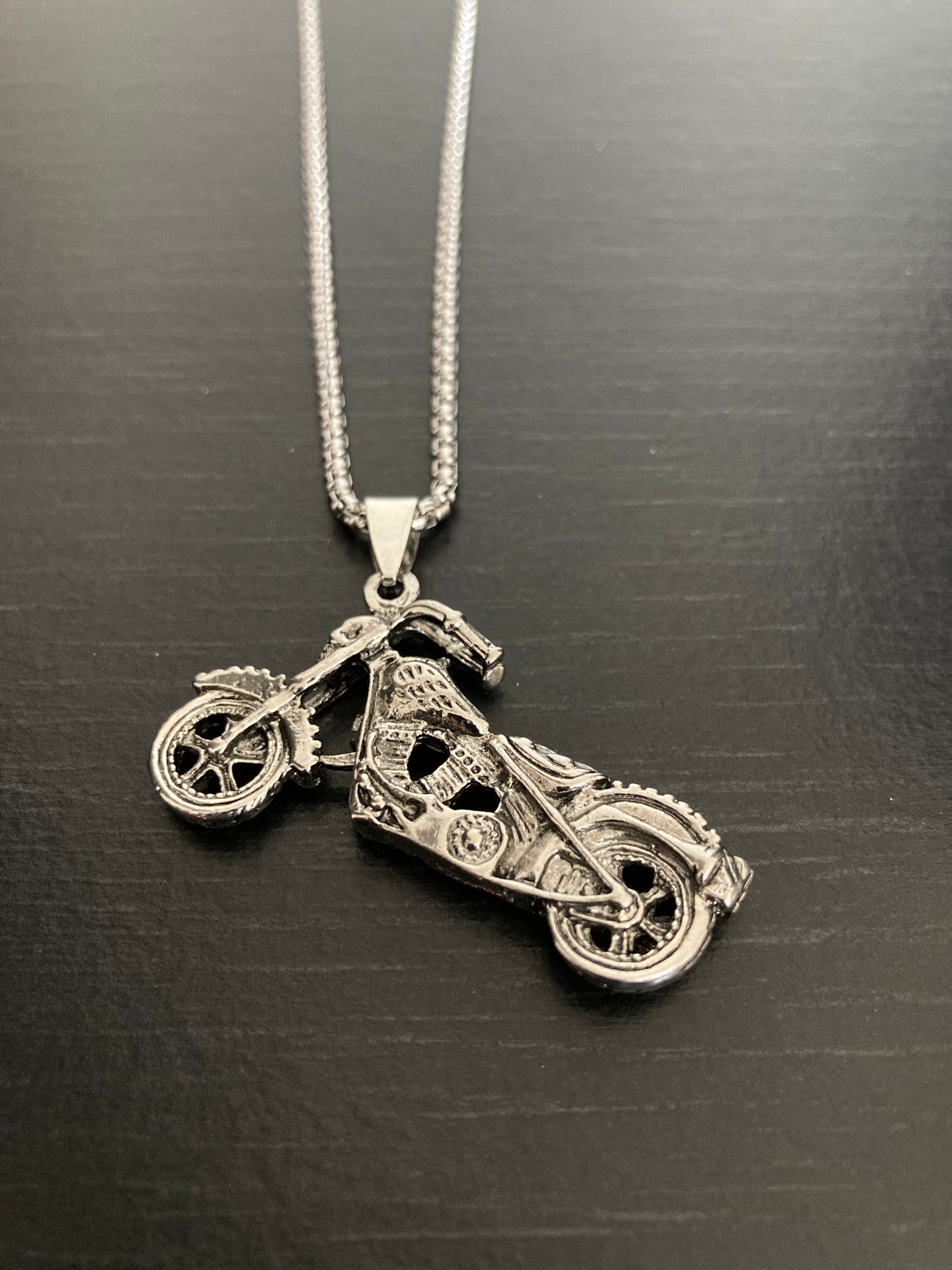 A chunky motorcycle pendant lies on a black surface attached to a chain. This is all silver in colour and made of quality stainless steel. The bike itself is incredibly detailed from the handlebars to the wheels and is beautifully made.