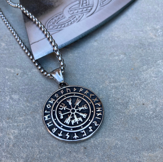 Circular pendant laying on the edge of a viking style axe. Raised pattern in the middle of the pendant and around the edges which is silver and black in colour.   Shiny in appearance and weighty in your hand. 