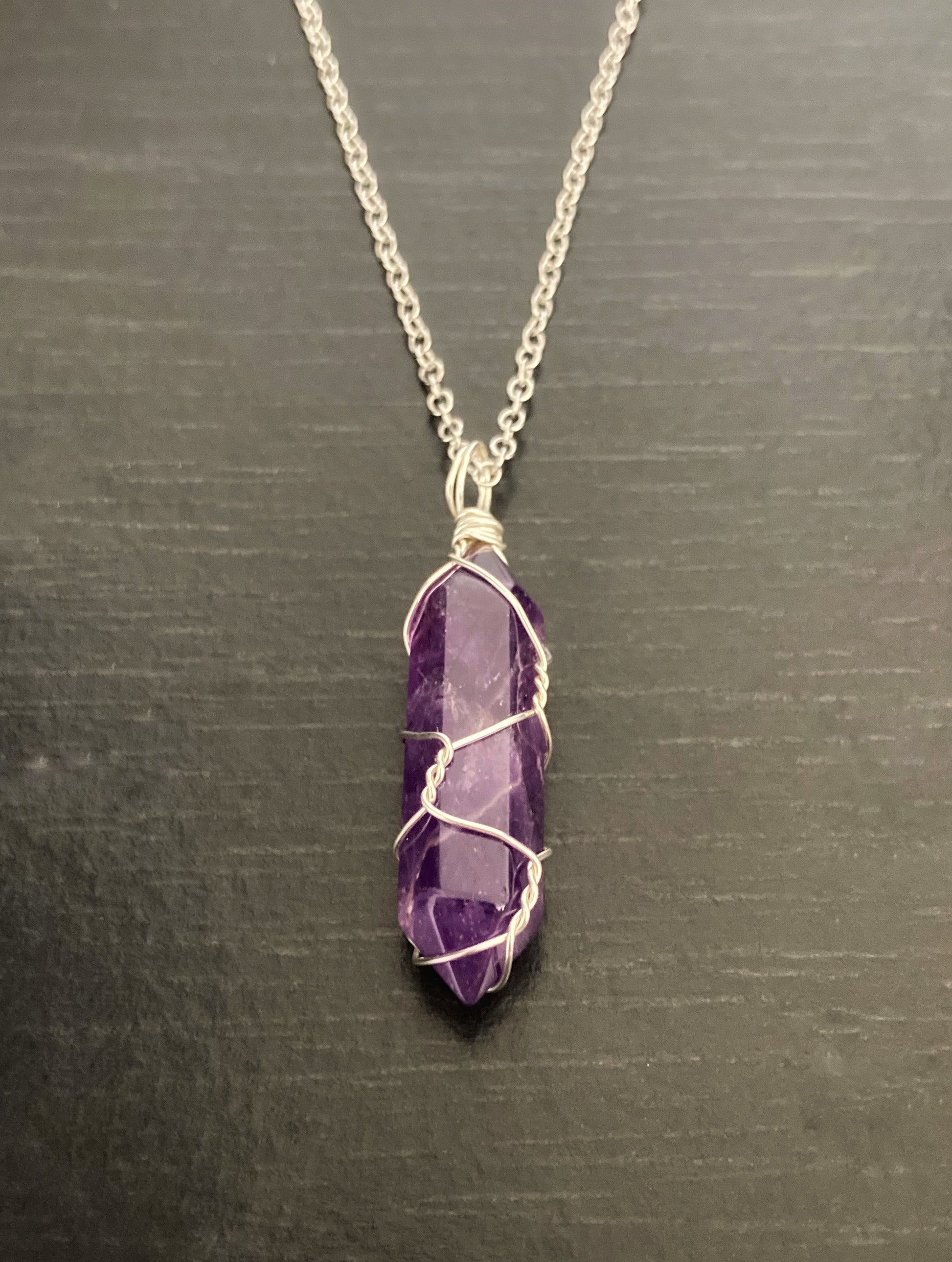 A front view of a crystal on a necklace chain. This is purple in colour with the chain being a lightweight silver stainless steel. The crystal has a shine to it and is wrapped in thin wire.