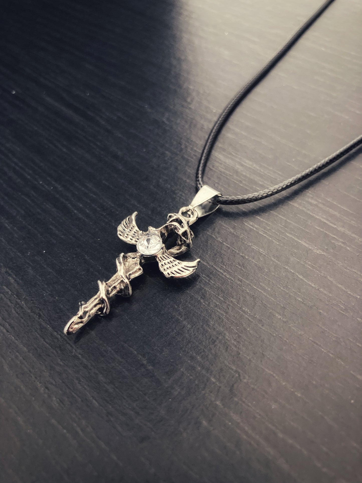 Lying at an angle is a dagger pendant with a serpent entwined around it. There are wings sticking out both sides and a clear glass stone attached to it as well. The whole thing is attached to a black corded necklace which is high quality.