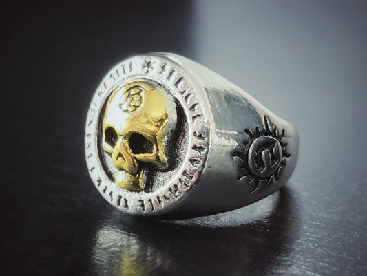 A close up view of a solid looking ring that features a gold coloured skull in the middle with patterning around the outside. The main part of it is silver in colour.