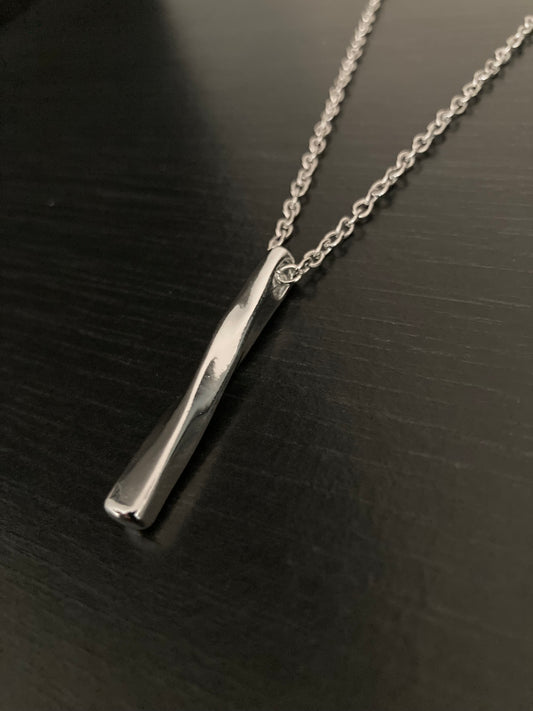 On a dark surface lies a necklace with a solid looking pendant which is rectangular in shape but twisted. The whole thing including the chain is a shiny silver colour.