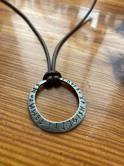 Sitting on a shiny wooden table is a stainless steel silver coloured circular hollow pendant. There are engravings of runes around the outside and it is attached to a brown cord necklace. The item is rustic and authentic looking and light to hold.