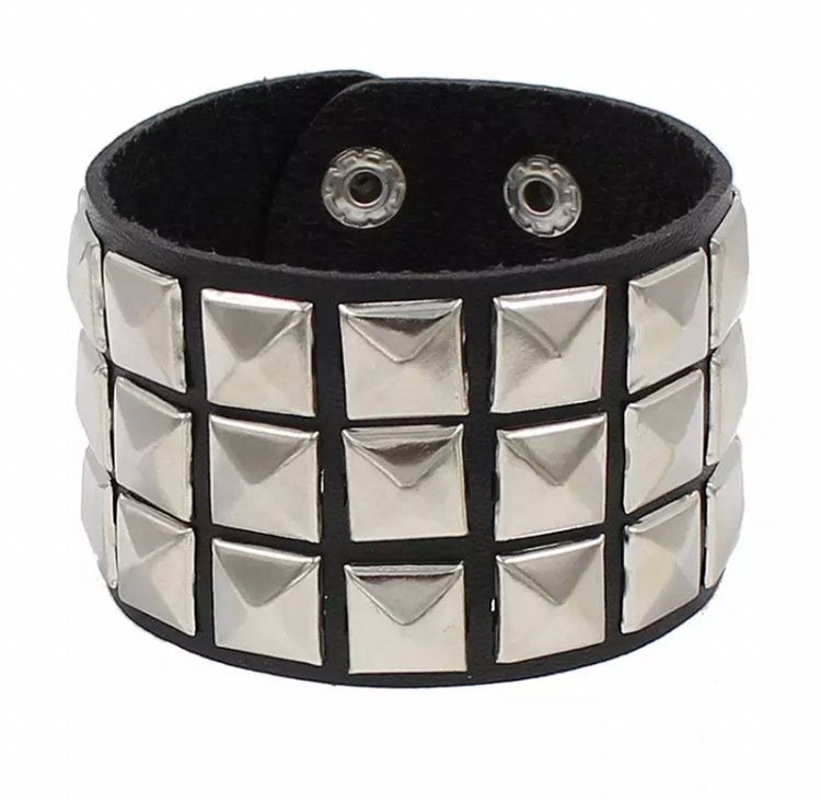 On a white background sits a black leather wide bracelet. Attached to the outside and covering the leather are silver coloured pyramid style shapes squares.