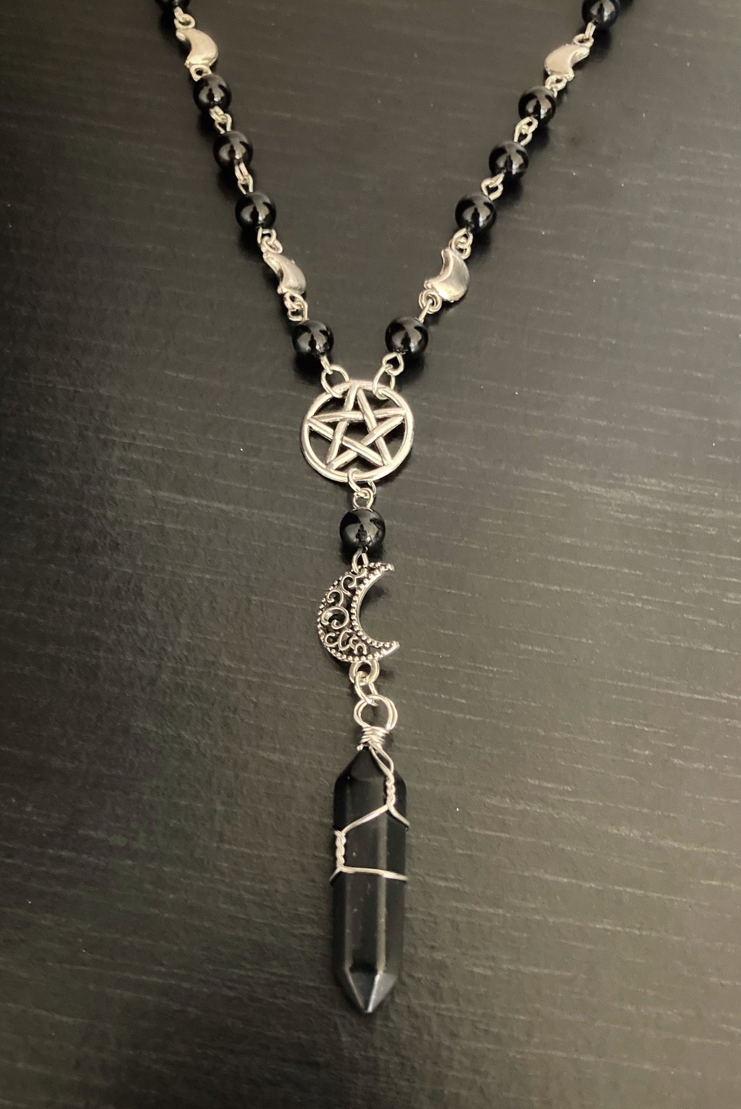 A view of a necklace that is both made up of chain and beads with a part at the front hanging down. This part has a pentagram in a circle with a crescent moon under it and at the bottom is a black onyx stone.