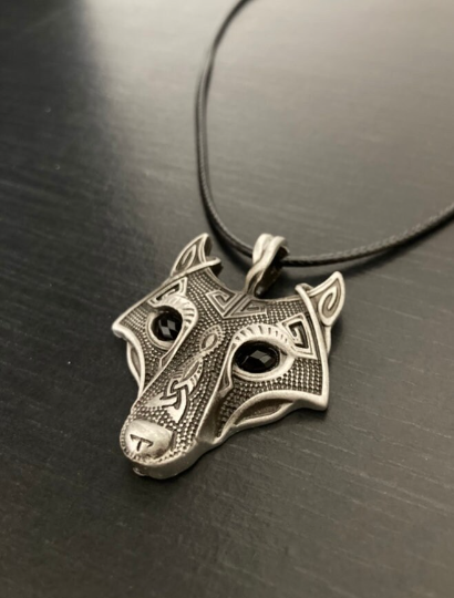 A pendant in the shape of a wolf's head is attached to a black cord necklace and sits on a black background. There are black stones for the eyes and very intricate engraved markings all over the face itself. It's silver in colour and shiny to look at
