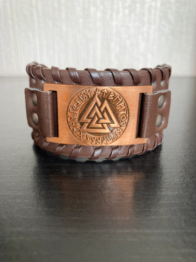 A brown leather bracelet sits on a plain background. This is a wide cuff style bracelet and the centrepiece is a metal square with a valknut and runes engraving on it. The leather itself is textured and has press studs either side of the main design.