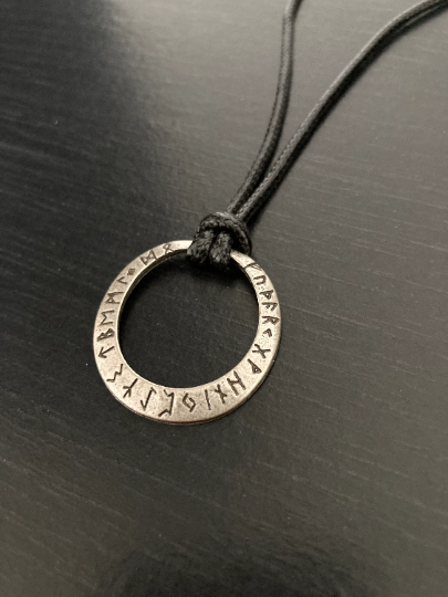 A rustic looking round silver coloured pendant is lying angled on a black surface. The detailing of the item is clear with runes that are etched on the outer part of it on display. The whole thing is lightweight to hold but still quality 
