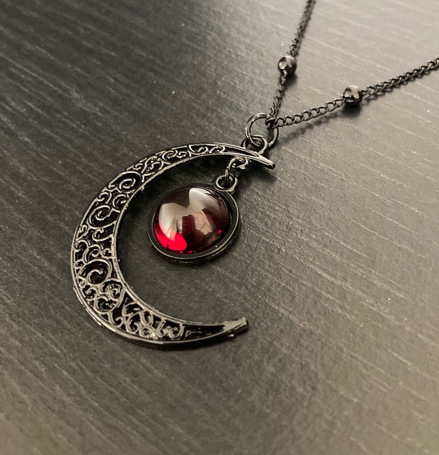 A necklace is lying on a dark surface. There is a crescent moon pendant with a dark red glass dome shape hanging from the tip of it. There is a thin chain and the whole thing is black in colour. The moon has patterns etched onto it.