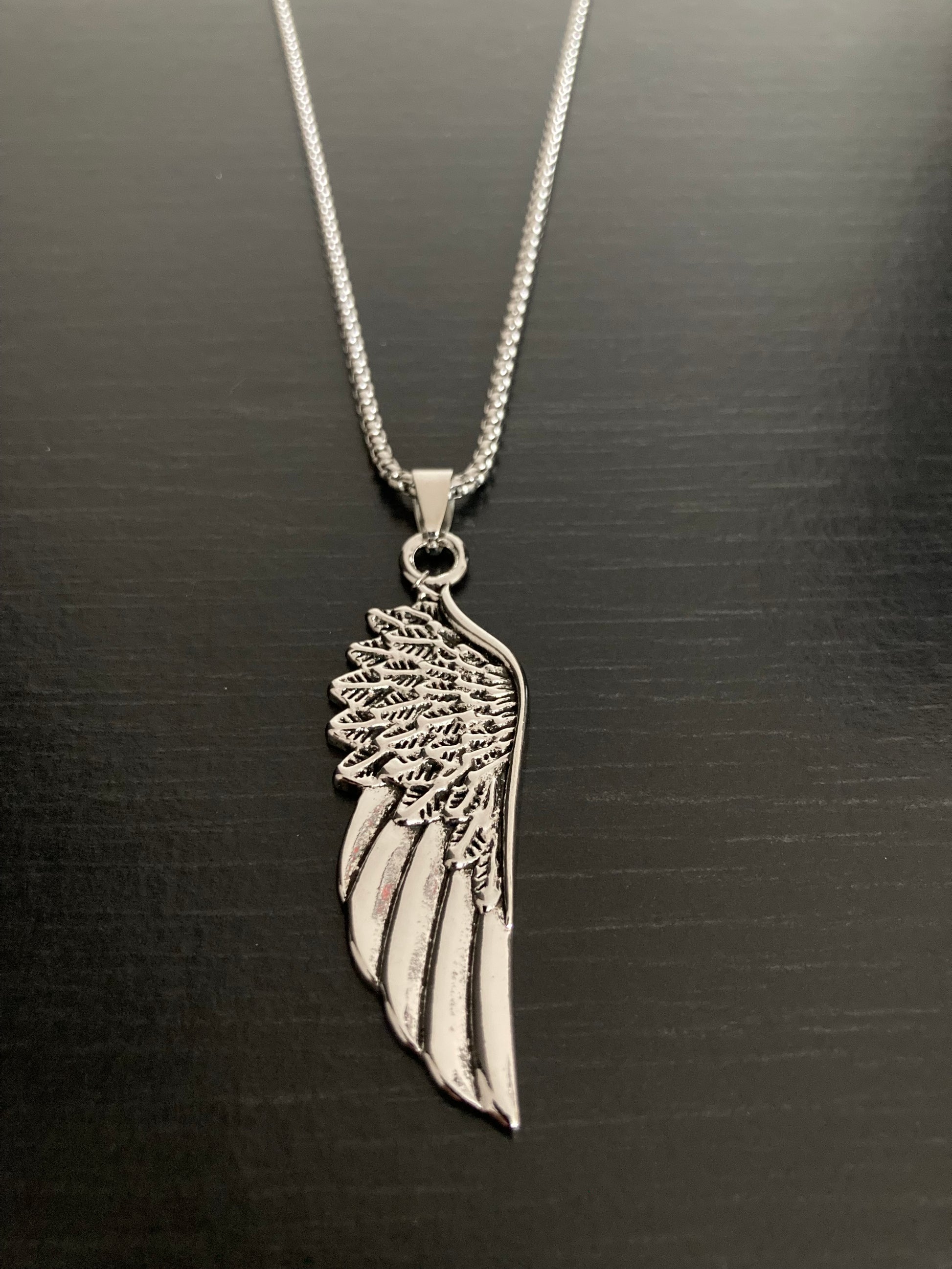 A close up view of a pendant in the shape of a wing. You can see the engraved markings on the item and if you touch it you can feel the detailing. The whole necklace is light in weight with the chain feeling strong.