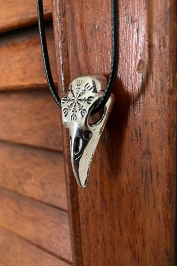 Hanging on a wooden door is a stainless steel raven skull pendant on a black cord necklace. It featuress a viking helm of awe design on the front part of the skull head and has hollwed out eyes. The cord has a metal clasp for fastening.