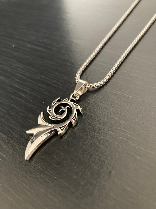 On a grainy black background sits a silver coloured chain and pendant which has a spiral design to it. There is the main spiral with a longer piece coming off of the end of it giving it a unique look and is shiny in appearance.