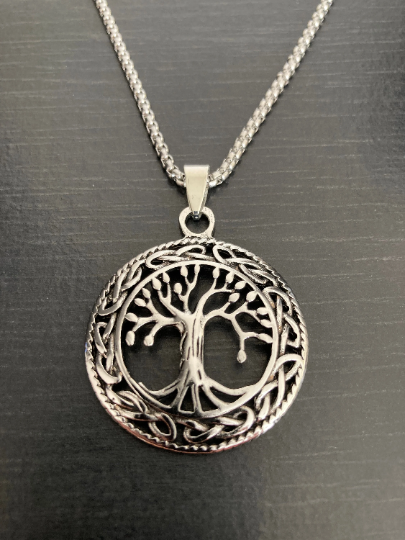 A necklace featuring the tree of life is sitting on a grainy black background. The pendant is round and silver in colour with the tree in the middle which is a cut out and a pattern that runs around the edge and is shiny to look at.