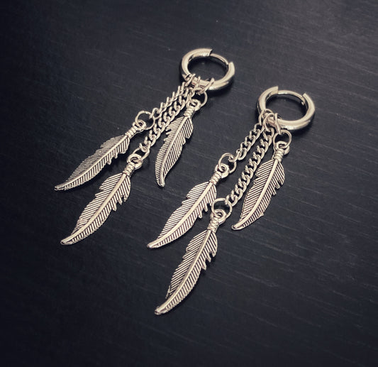 On a black surface lies a pair of hoop earrings. These are fairly small hoops and each one features 3 silver coloured feathers hanging from chains. These are different lengths so the feathers arent all the same height.