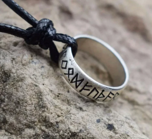A silver coloured ring attached to a black cord necklace sits at an angle against a rock. The outer part of the ring is engraved in black and features rune symbols all around the outside of it. 