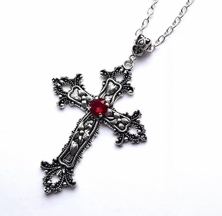 On a white background sits a gothic looking cross that features a red stone in the middle of it. There are flower designs etched onto the cross and it is attached to a chain necklace with the whole thing being a silver colour.