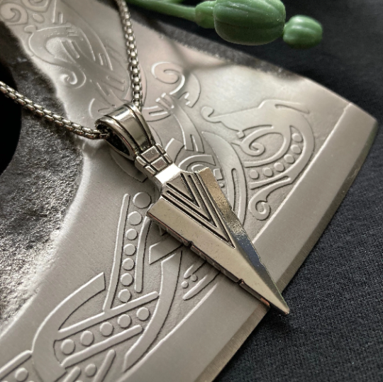 Lying on an axe blade is a silver coloured arrow head pendant and chain. This is shiny in appearance with v shaped markings on it. Lightweight to hold but still of high quality this pendant is made from stainless steel.