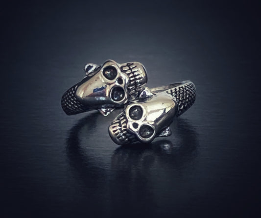 On a dark surface lies a silver coloured ring. There are two skull heads which sit side by side making the ring chunky and slightly twisted in design. 