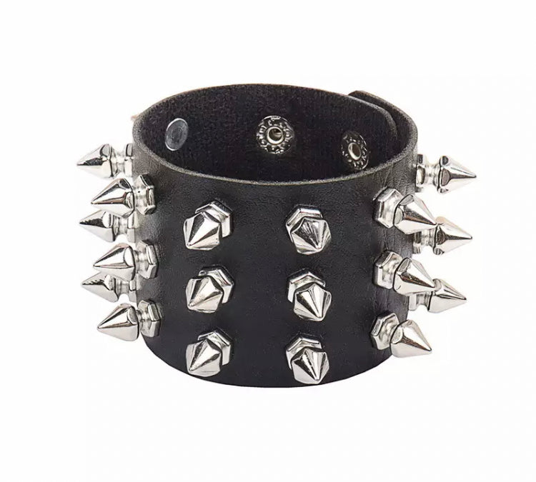 A black leather wide bracelet is on a white background. Attached to the outside circumference are silver coloured spikes. There are 3 per row going around it.