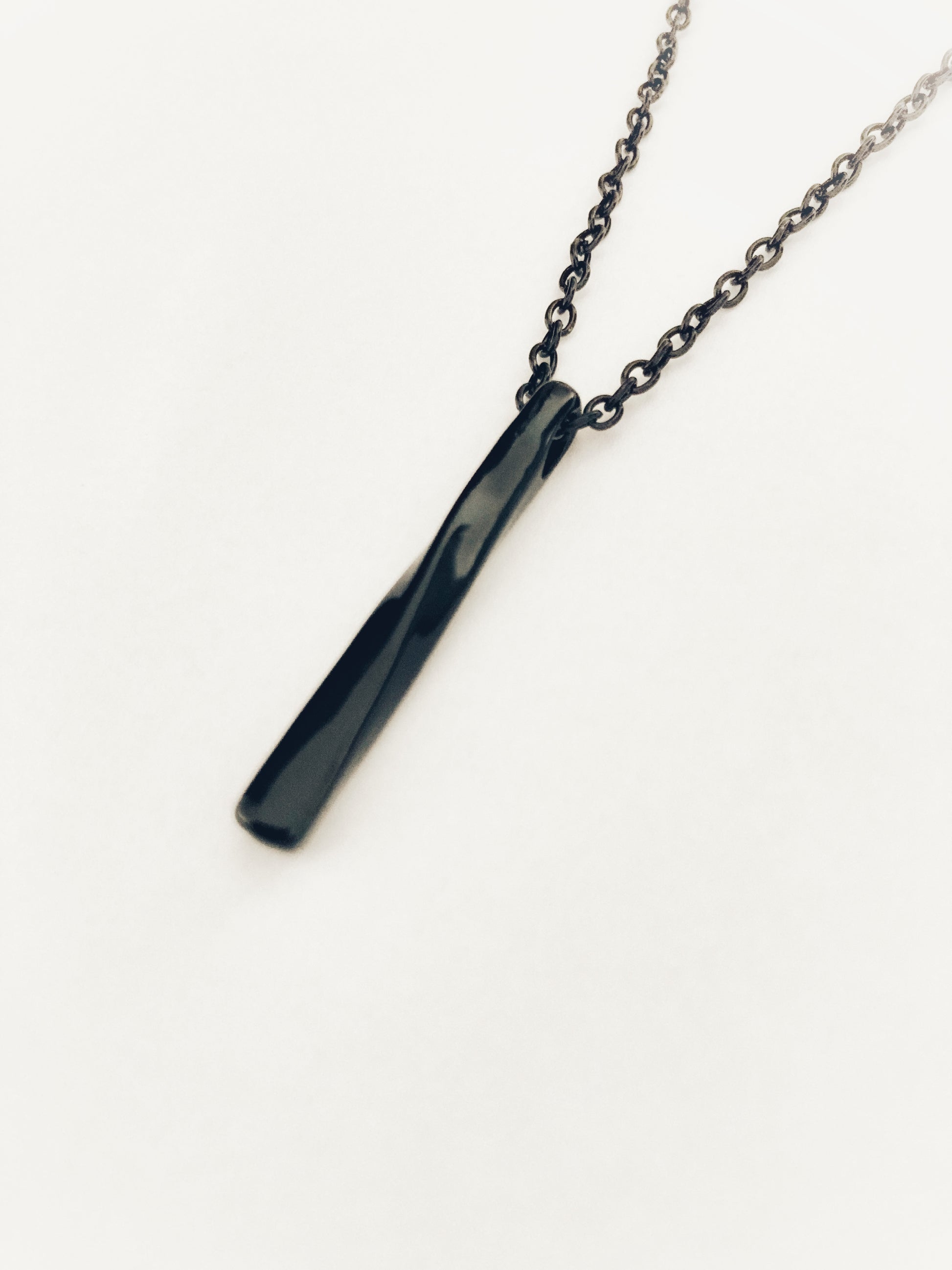 On a white background hangs a pendant that is black and is in the shape of a bar. It is twisted and is solid and is attached to a black chain.