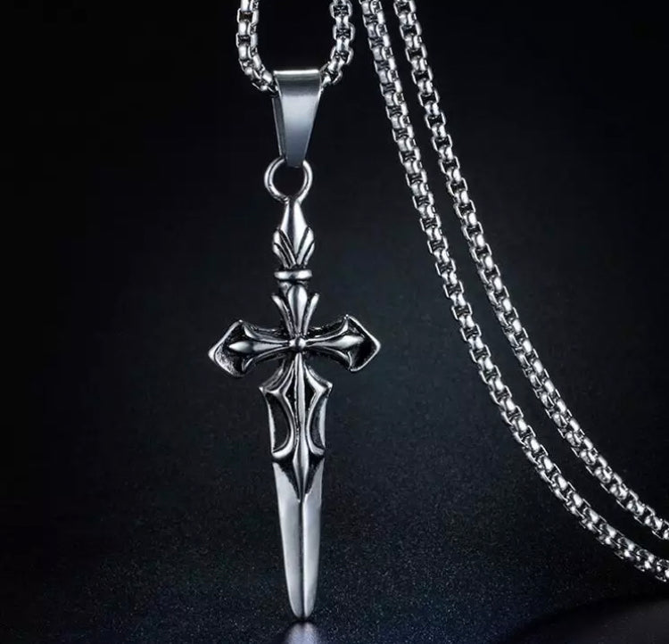 On a black background hangs a silver coloured pendant in the shape of a sword. The hilt of the sword is in a cross with engraved patterning on it. Attached is a sturdy chain in the same colouring.