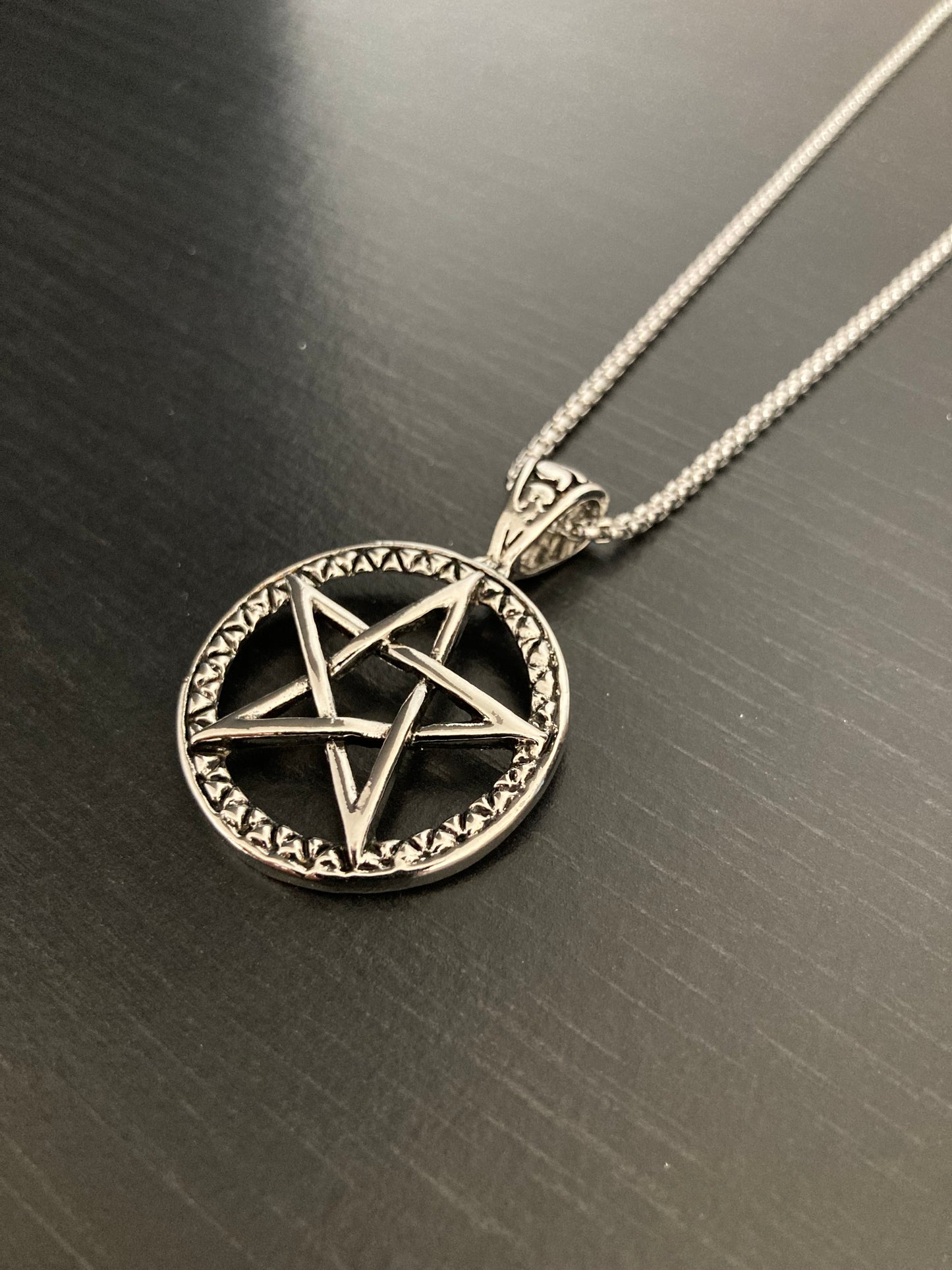 On a black background sits a shiny silver coloured necklace consisting of a chain and a circular pendant with a pentagram in the middle of it. This is a cut out design and the five points touch the outer circle of the pendant.