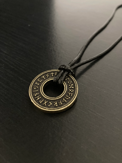 A round pendant that looks like a coin with a hole in the middle is attached to a black cord necklace. The circular shape is a rustic gold colour with runes engraved on it on the outer ring.