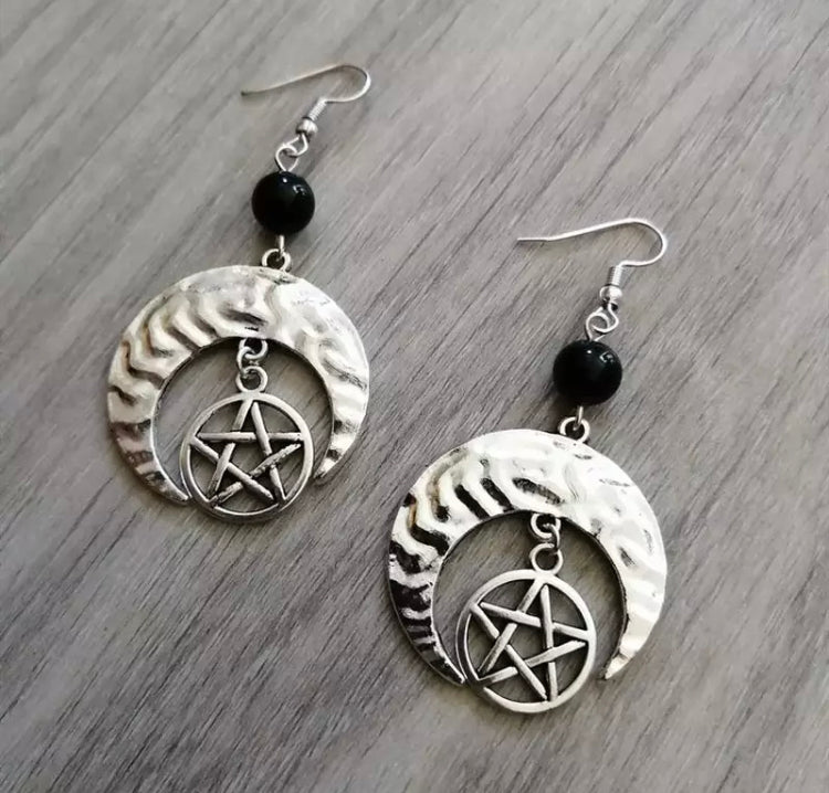 A pair of dangly earrings featuring a crescent shaped moon and a pentagram that hangs from the inner part of that moon. There is a black bead between the main part of the earring and the top part. These are silver in colour and shinny to look at.