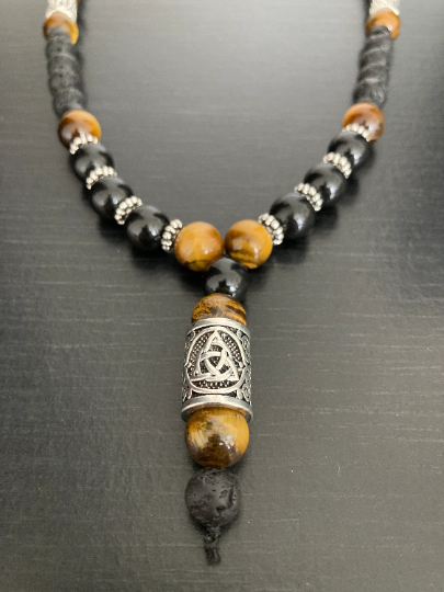 A beaded necklaces sits on view that is round with a section that hangs down at the front. This part featrures a metal silver coloured rune with a triquetra design engraved onto it. The beads around it are tigers eye and are yellow/gold in colour. 