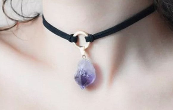 A choker style necklace is around someones neck.  With a black material cord that is tied 2 ends to a silver colour metal ring there is the a purple crystal hanging down from that ring.