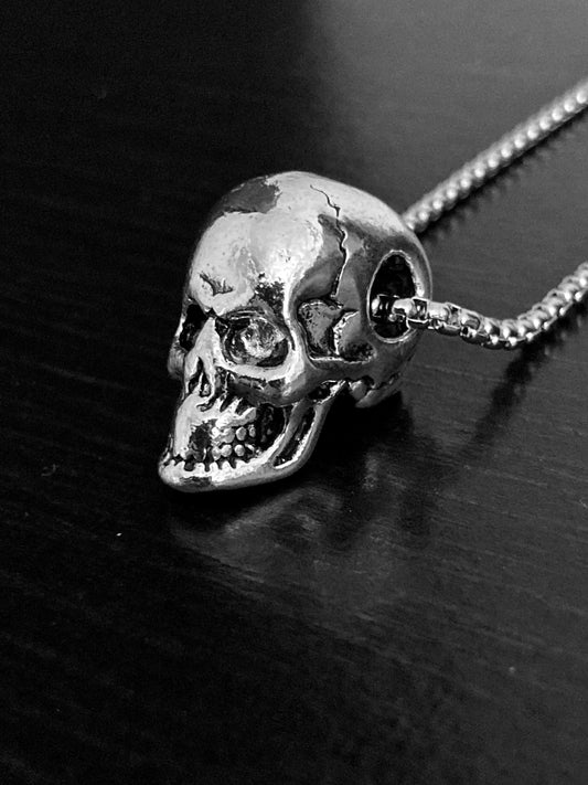 A side view of a solid skull head pendant. It is silver in  colour as is the sturdy chain and is stainless steel. The skull has detailed features including sunken eyes, a wide grimace and cracks in the skull. This is a chucky but not too weighty item