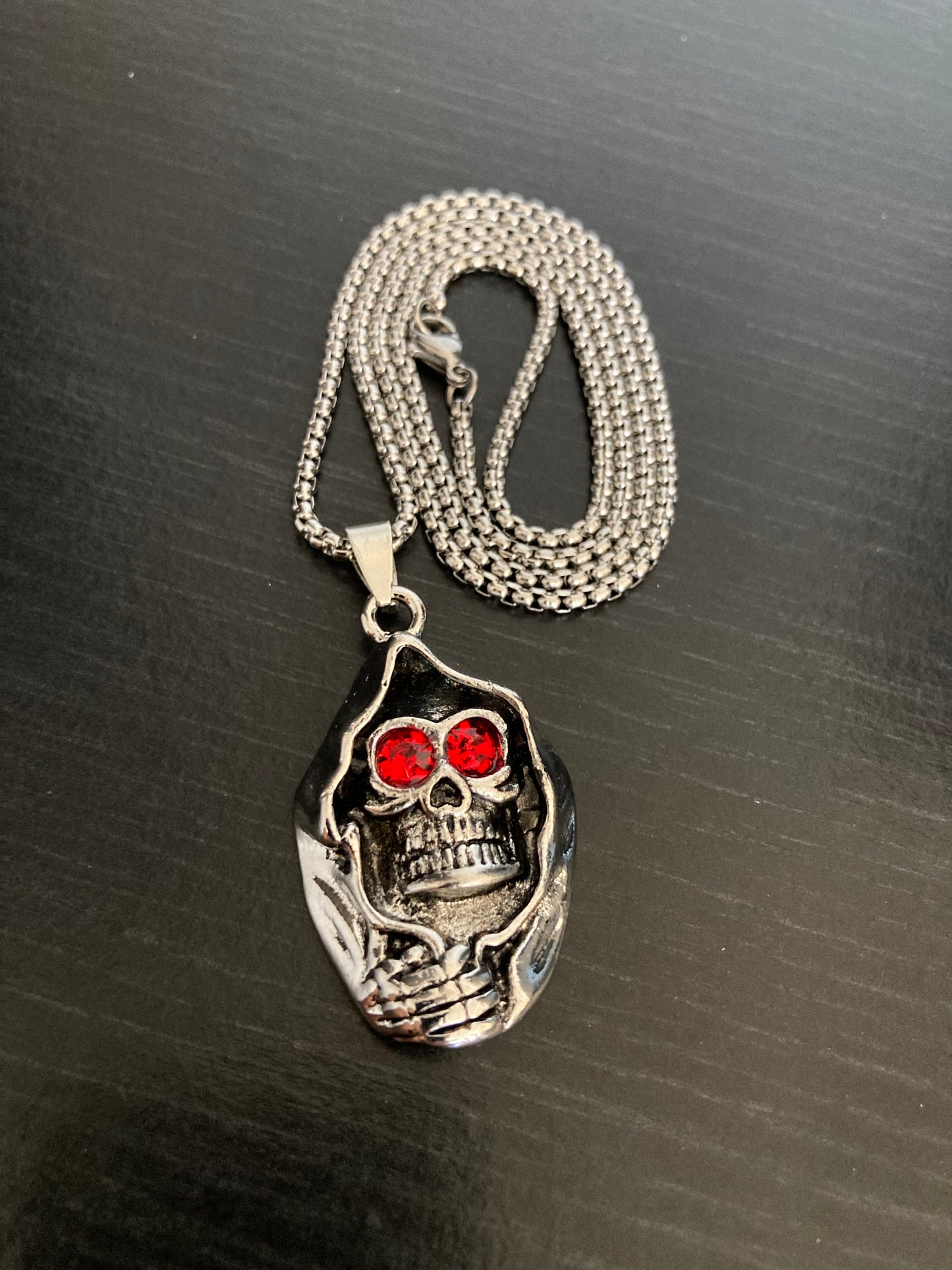 A silver coloured chain with metal clasp is curled on a black surface. Attached to it is a cloaked skull head pendant which has red stone eyes and a grimace showing its teeth. The whole item is stainless steel and is solid in quality. 